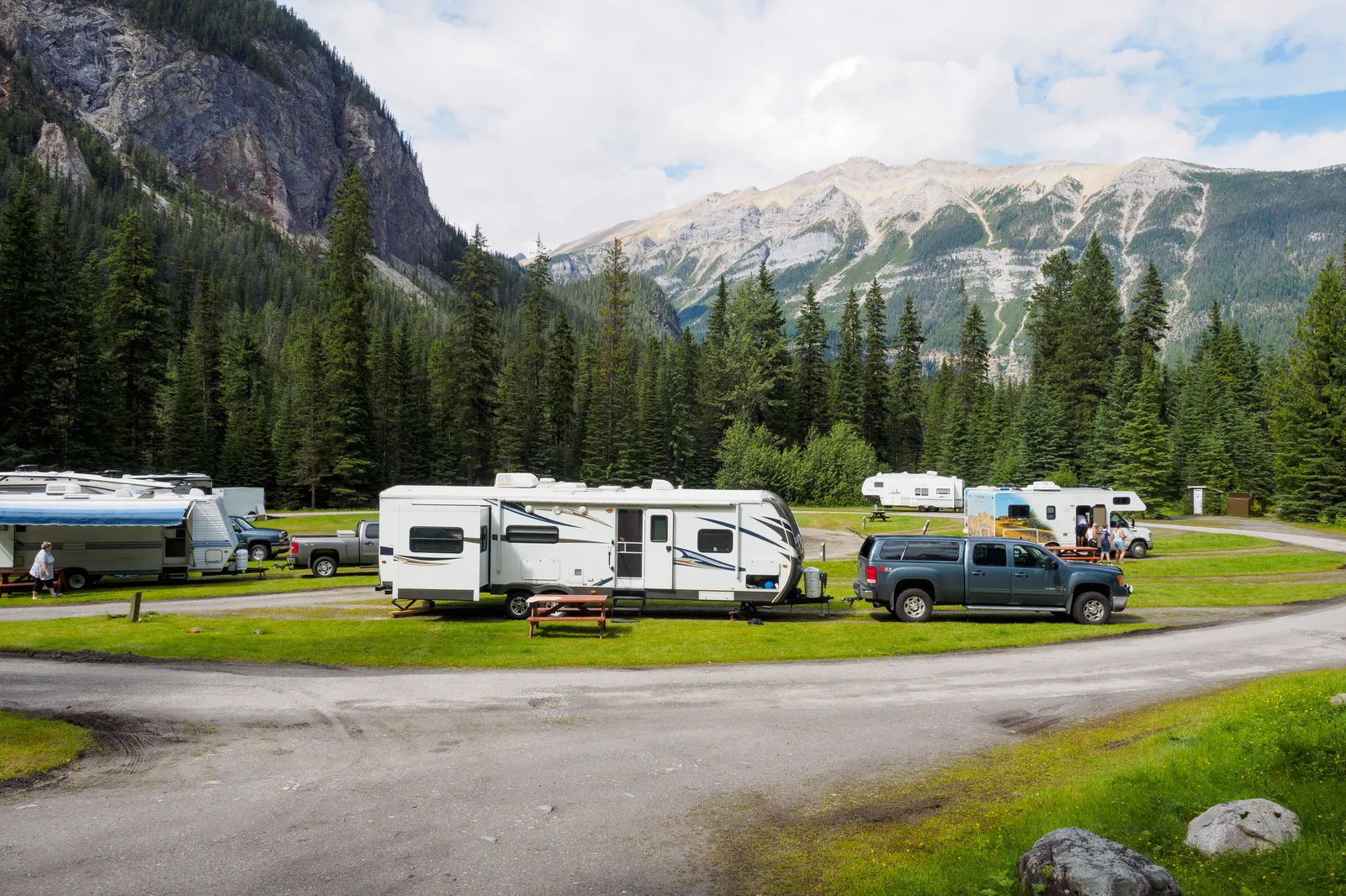 Travel trailers and trucks parked on numbered sites along a paved campground loop below a mountain ridge