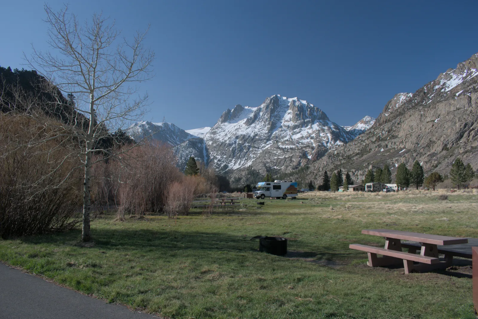 An empty campsite with a picnic table and fire ring at a developed campground below snowy peaks, a motorhome parked in the distance