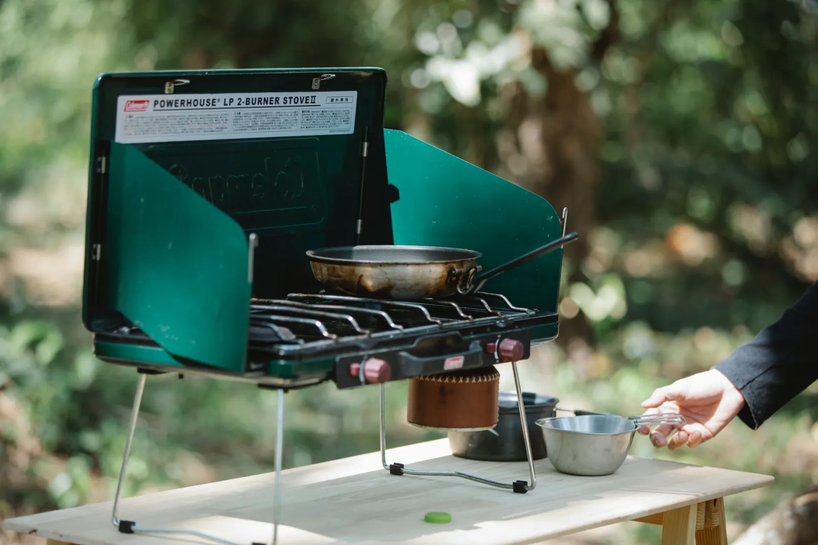 A person cooking with a pan on a portable gas burner in a forest