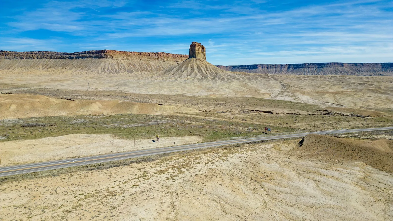 A two-lane highway crossing open desert badlands below an eroded butte and a long mesa rim