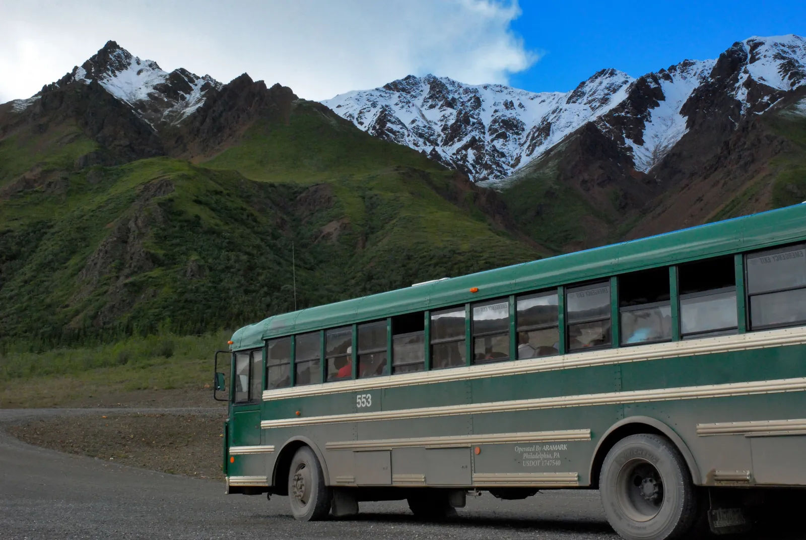 A green park transit bus on a gravel road below snow-capped mountains