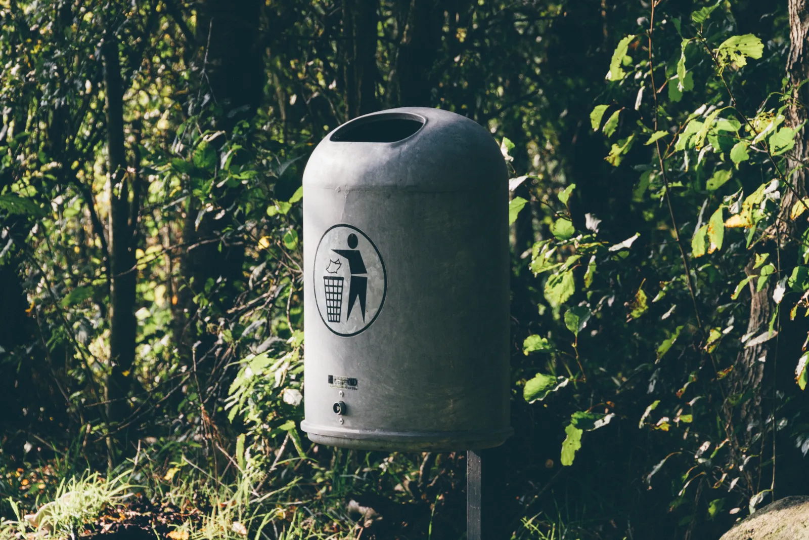 A gray metal trash can on a post among green trees, marked with a person-dropping-litter symbol