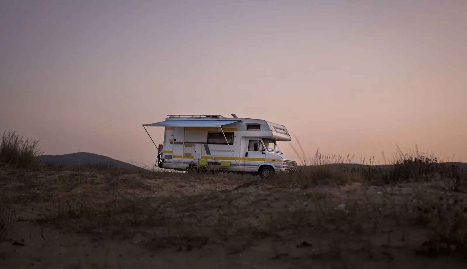 A motorhome with its awning out parked alone on open dry ground at dusk