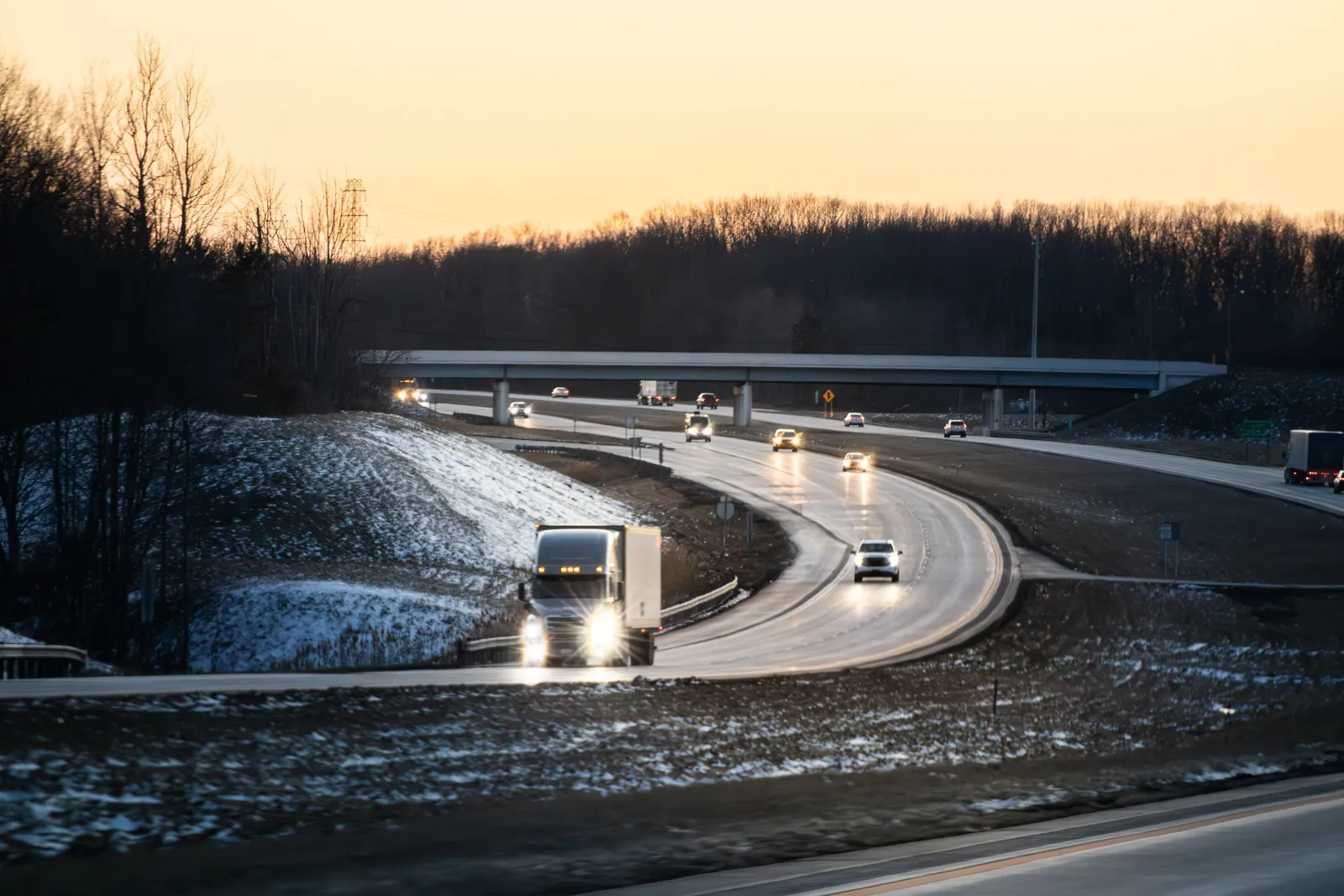 Cars and a semi truck on a divided highway at dusk with snow on the roadside banks