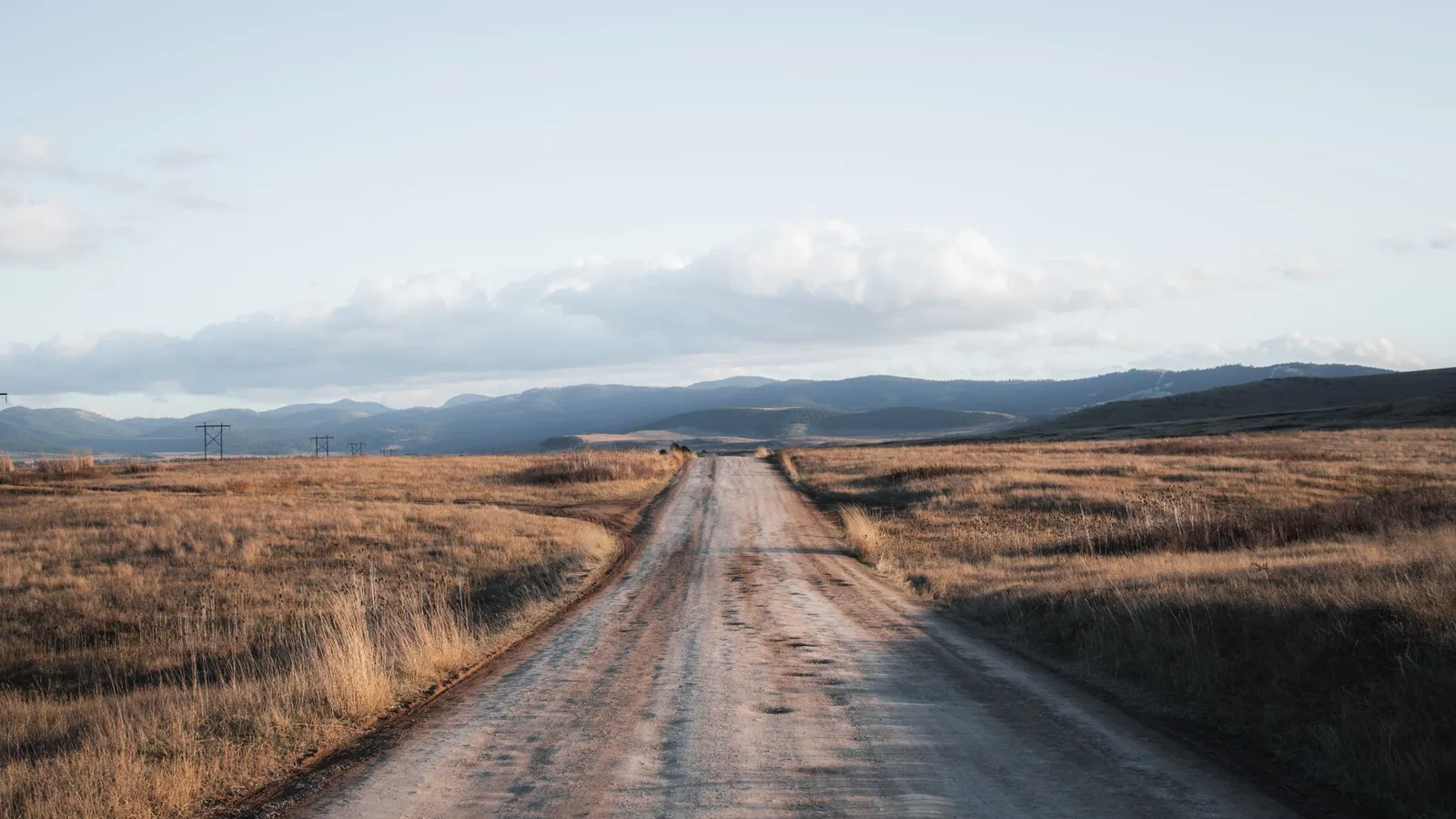 A gravel road running straight through dry golden grassland toward low forested hills