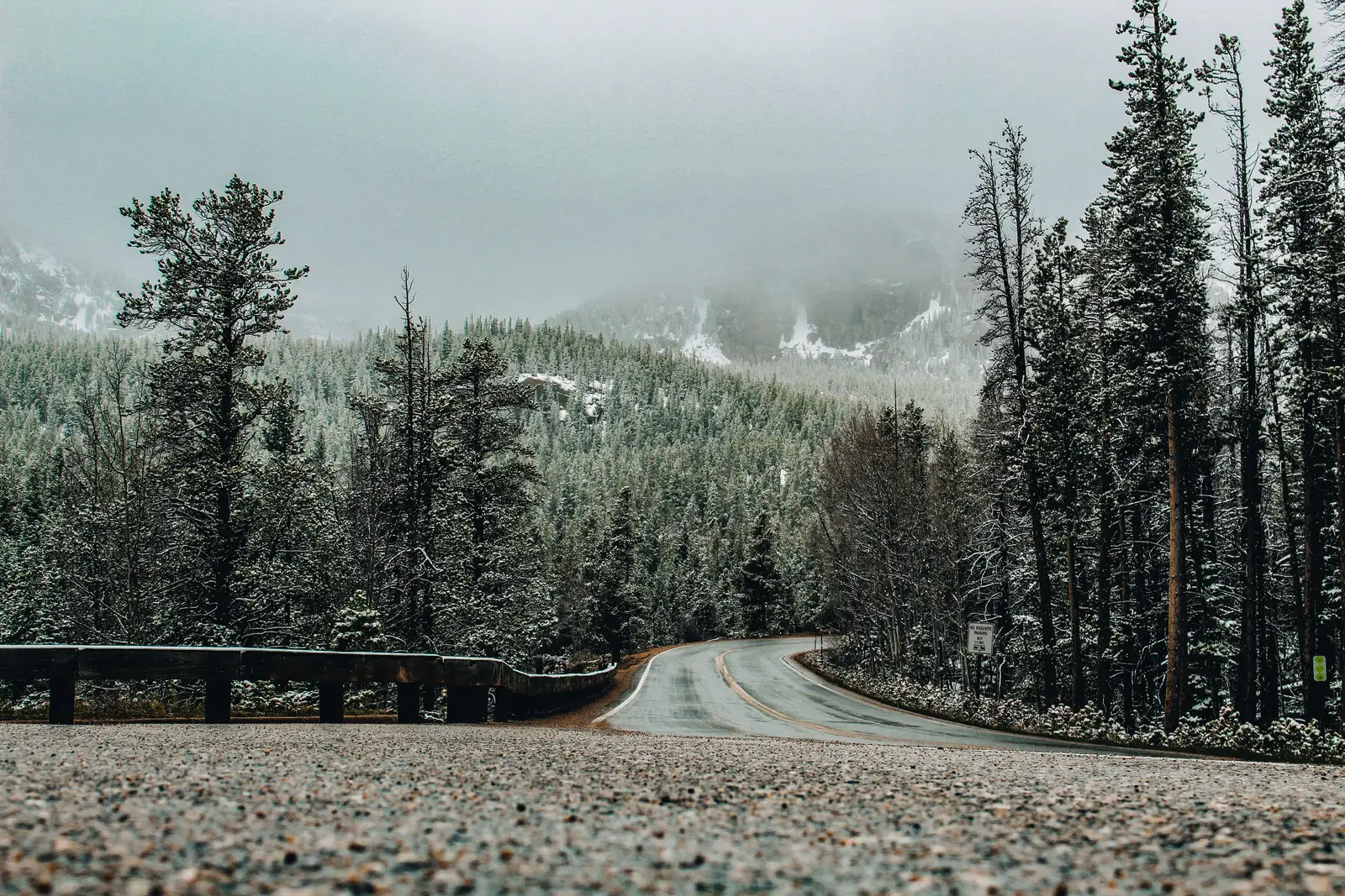 A two-lane road curving through snow-dusted evergreen forest below cloud-covered mountains
