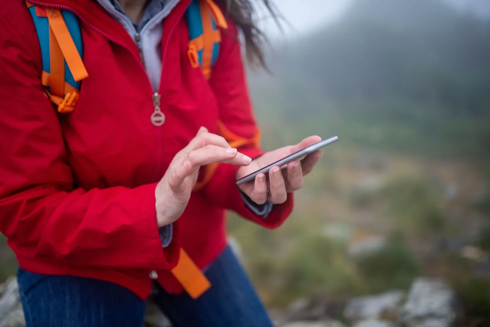 A hiker in a red jacket using a smartphone on a rocky slope in fog
