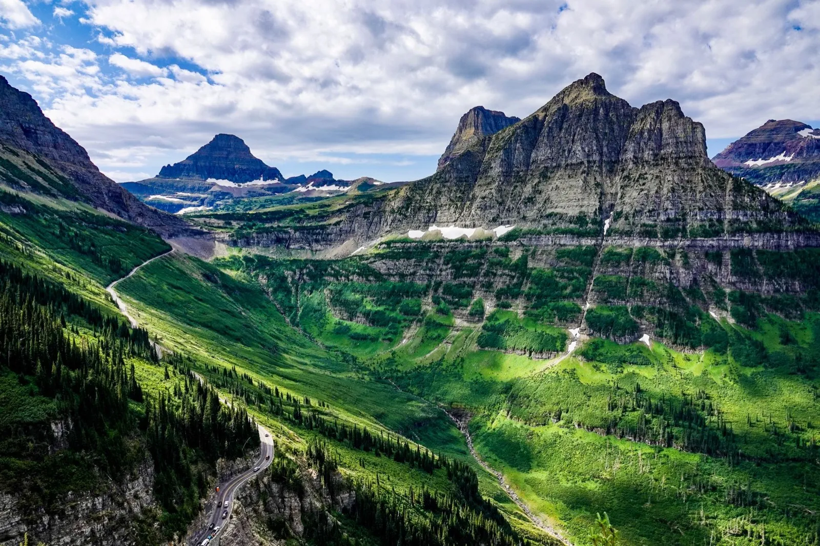A highway curving along a green mountainside below rocky peaks in Glacier National Park
