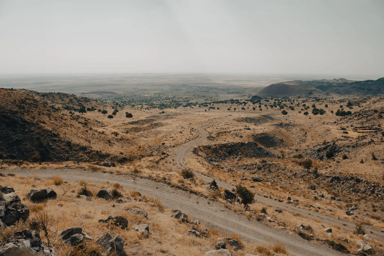 A dirt track winding down through dry rocky hills toward a hazy open basin