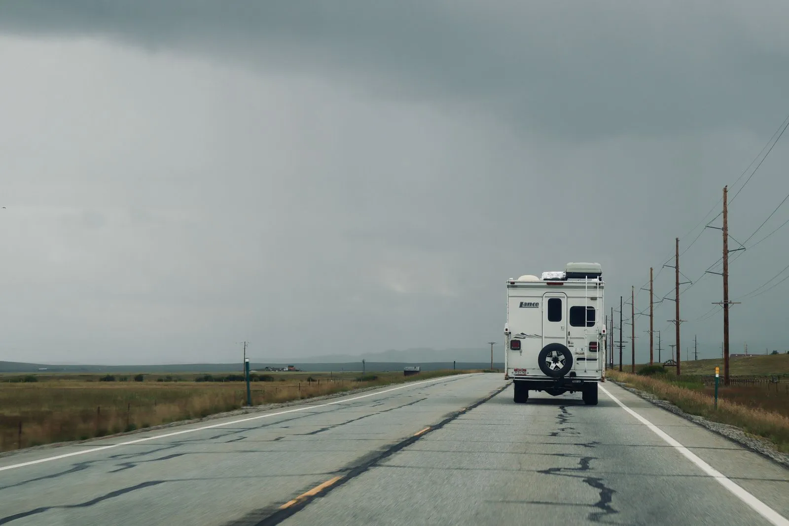 A pickup truck camper driving away on a two-lane highway under heavy gray rain clouds