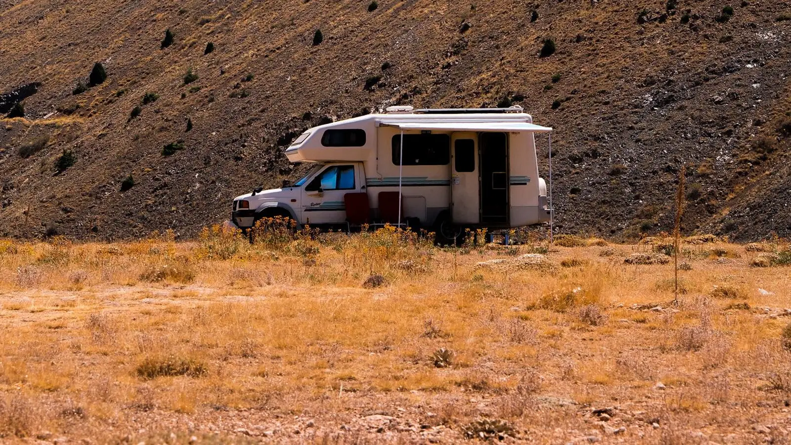 A small motorhome parked alone in dry grass below a bare hillside dotted with junipers