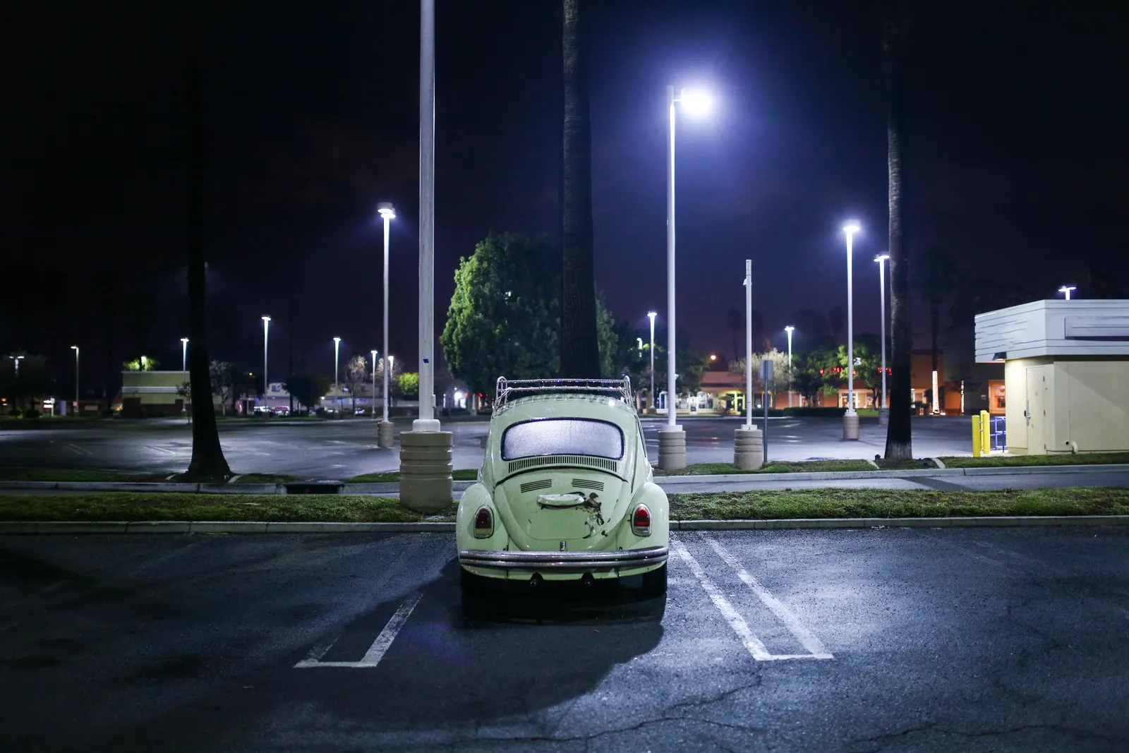 A single pale green car parked alone in an empty parking lot under bright lights at night
