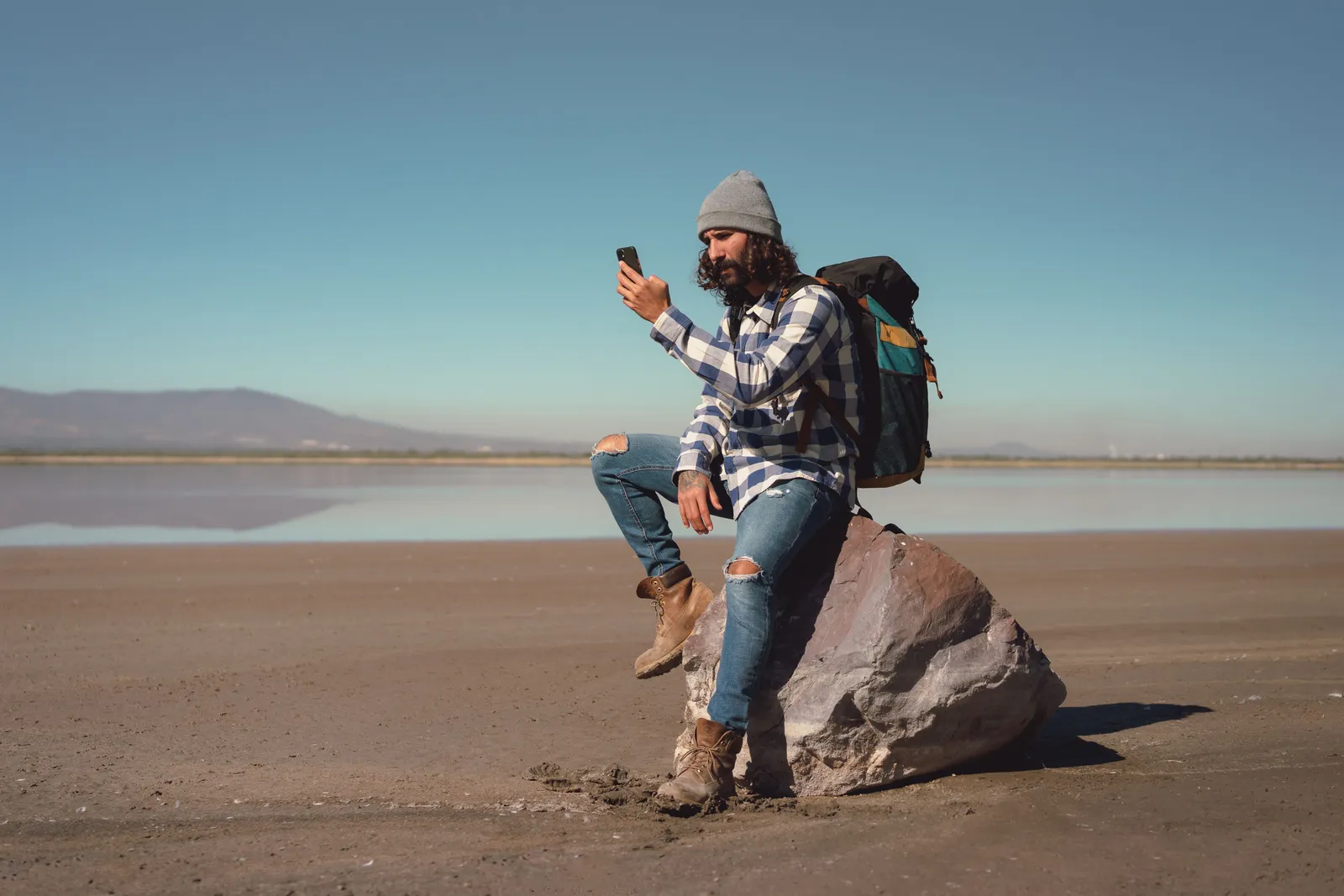 Hiker with a backpack sitting on a rock in open terrain, looking at a phone under clear sky