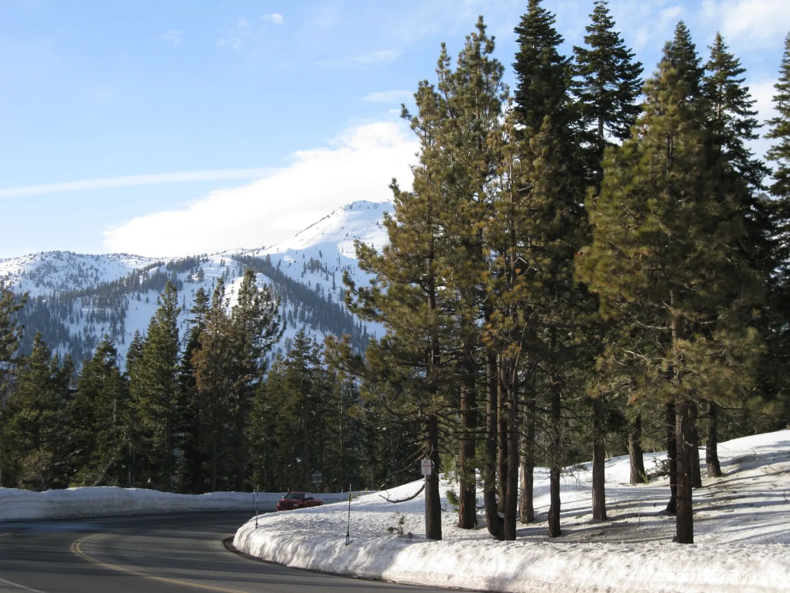 A plowed two-lane mountain road curving past snowbanks and pines below a snow-covered ridge