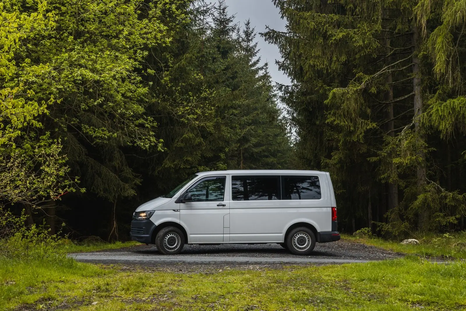 A plain white van parked on a gravel pullout at the edge of a conifer forest