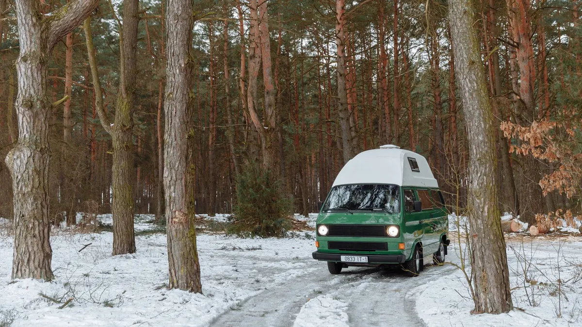 A green high-top camper van parked on a snow-covered track in a pine forest