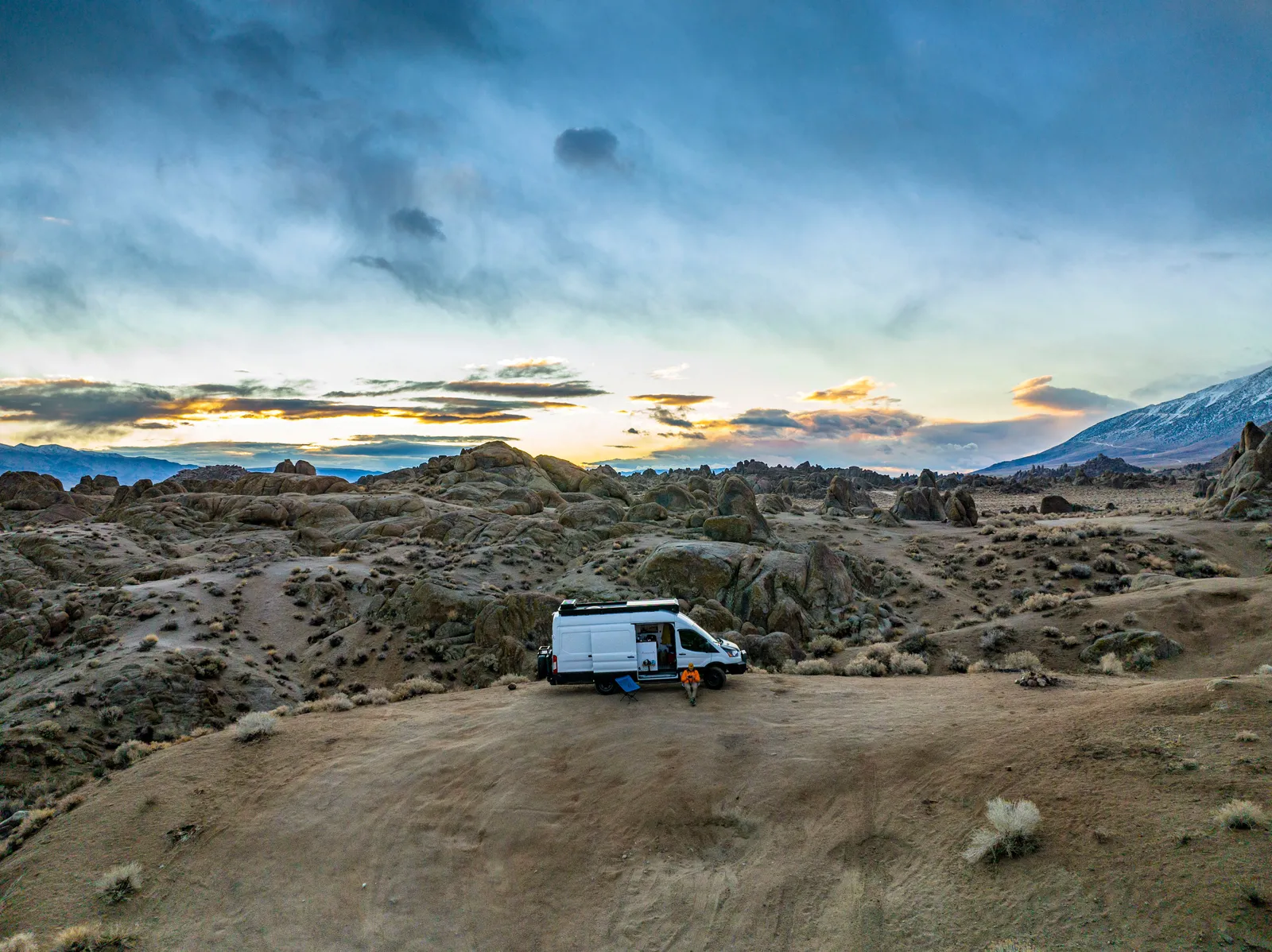 A camper van parked among desert boulders at dusk with a portable solar panel set out on the ground
