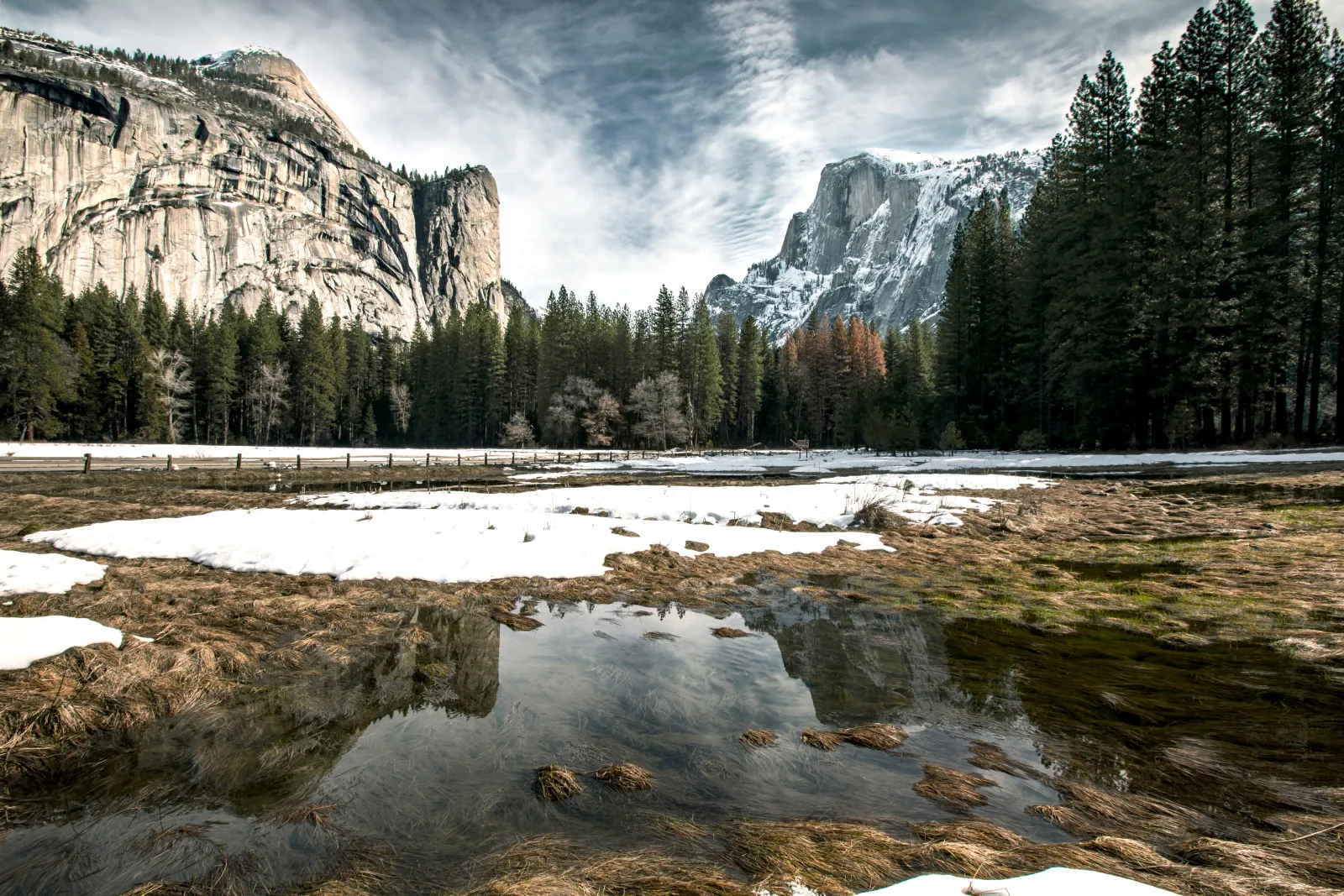 Granite cliffs and a snowy peak above a meadow with patches of snow and standing water